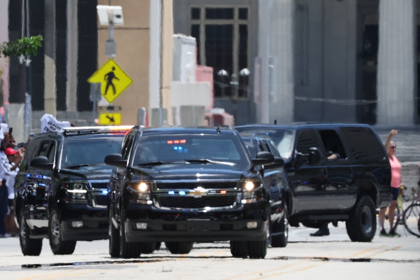 A motorcade of cars gathered on a road with a person walking in the background.