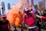 A young Israeli man wearing a bright pink T-shirt and a snare drum holds an orange smoke canister during a street protest.