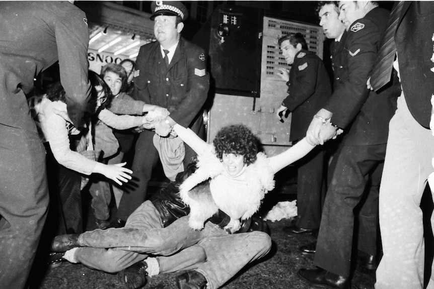 A woman is photographed in black-and-white on the ground as police officers grab one arm and protesters grab the other.