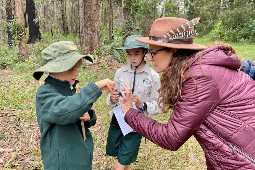 A woman wearing a hat and puffer jacket shows a boy in a green uniform and hat a leaf with another male student watches on