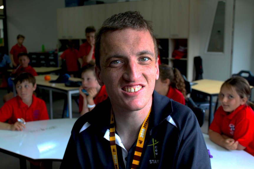 A young man smiles with school students in the background behind him in the classroom.