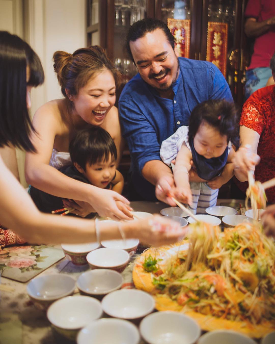 Adam Liaw, smiling and holding a small child, stands with others adults around a table covered in bowls and food.