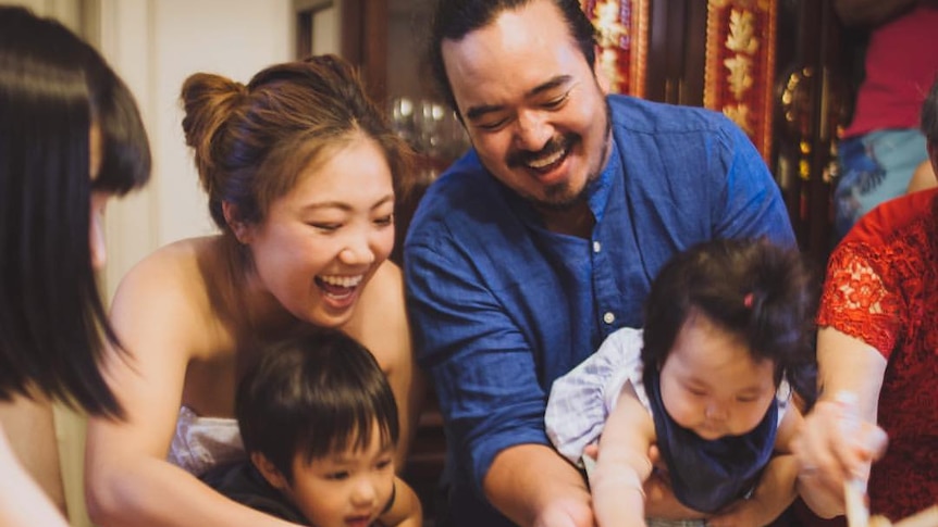 Adam Liaw, smiling and holding a small child, stands with others adults around a table covered in bowls and food.