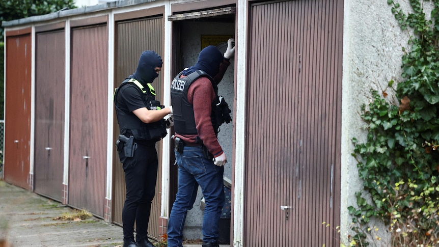 Two plain clothes police in bullet proof vests stand in the door of a shed. 