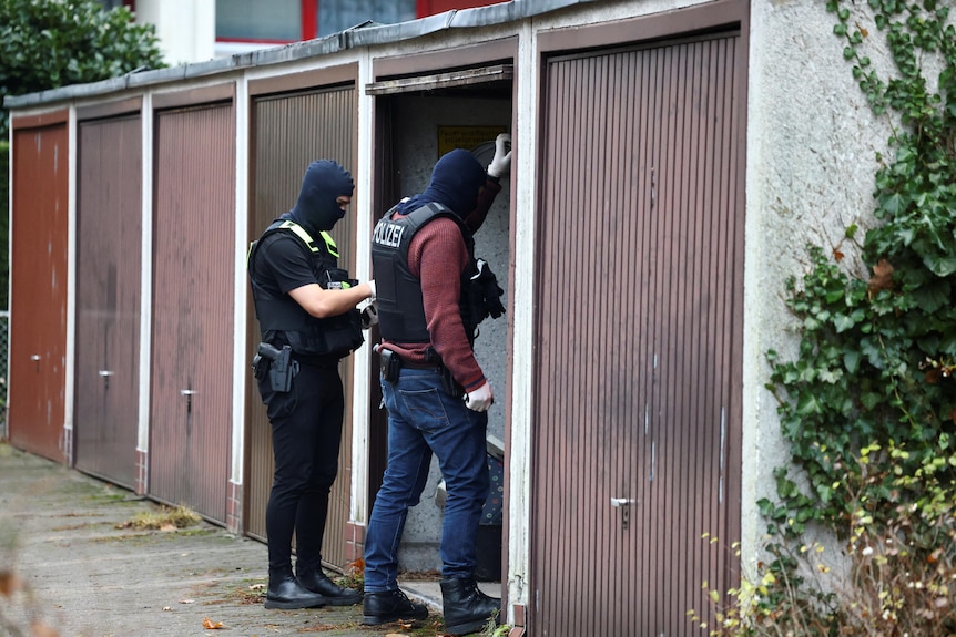 Two plain clothes police in bullet proof vests stand in the door of a shed. 