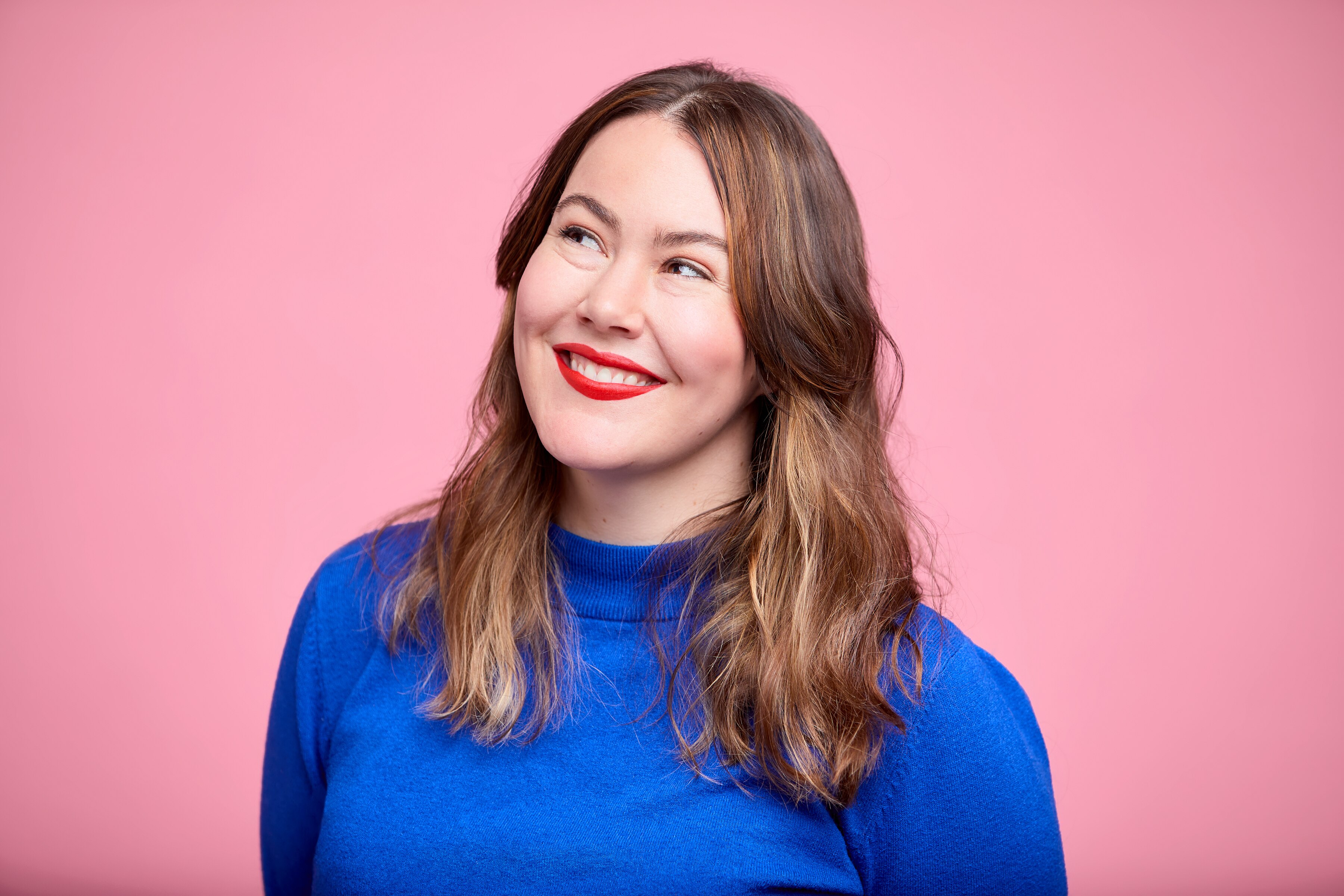 Writer Madeleine Dore smiles while looking off camera, wearing a blue turtleneck and red lipstick, in front of a pink backdrop.