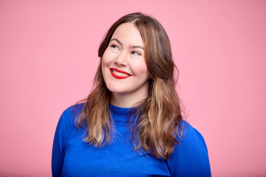 Writer Madeleine Dore smiles while looking off camera, wearing a blue turtleneck and red lipstick, in front of a pink backdrop.