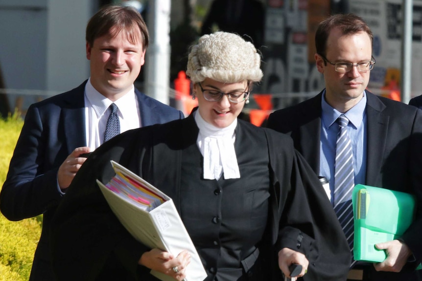 Two white men with a woman in a legal wig and gown in the sunlight mid-walk.