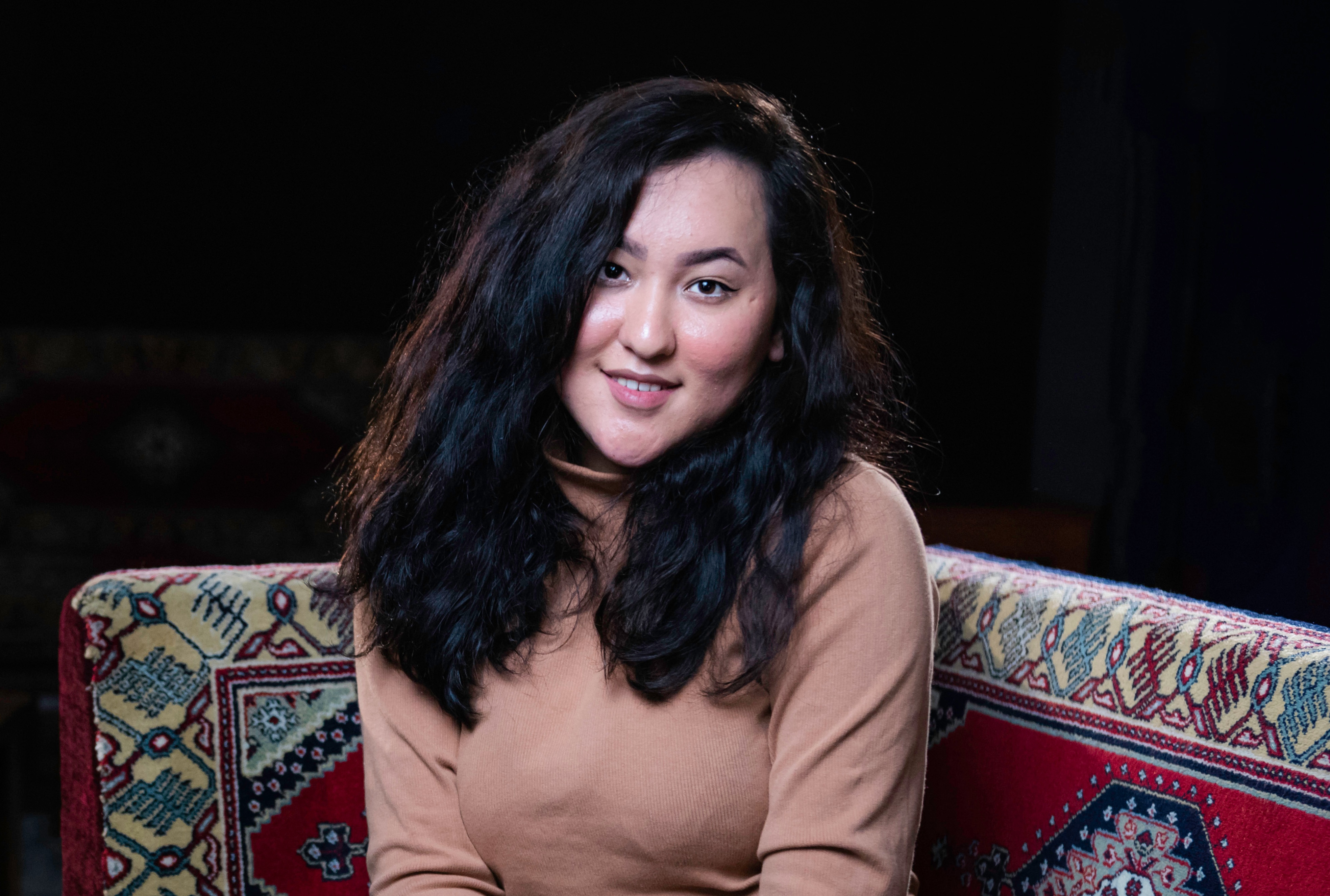 A young Afghan woman smiles as she looks into the camera lens. She is sitting on an embroidered lounge.