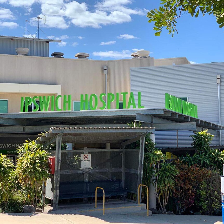 Entrance to Ipswich Hospital, flanked by trees. There is a yellow sign to one side, which reads "COVID-19 Fever Clinic".