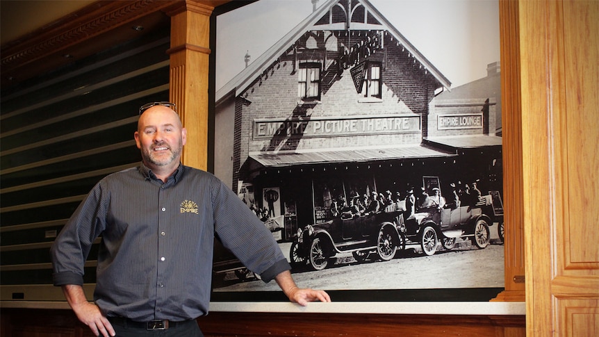 Empire Cinema owner Gerard Aitken stands in the foyer of the cinema next to a historical photograph.