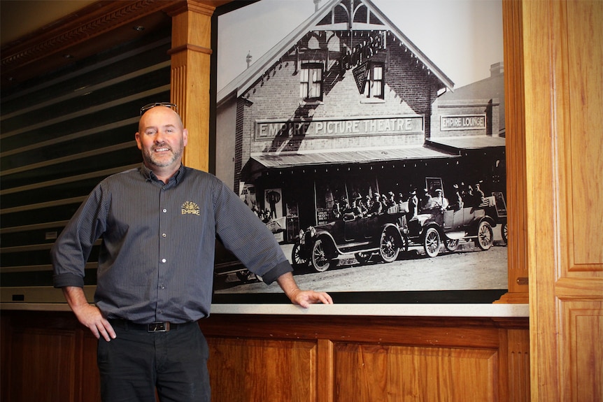 A smiling man in a dark work uniform stands next to a large historic photograph.
