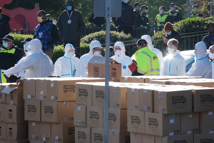 Men dressed in white infection control suits stand behind a large stack of boxes.