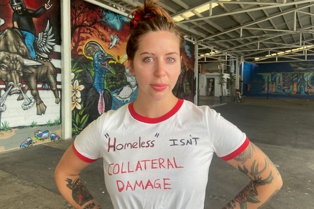 A woman standing with her hands on her hips. her shirt says 'homelssness is's collateral'.  