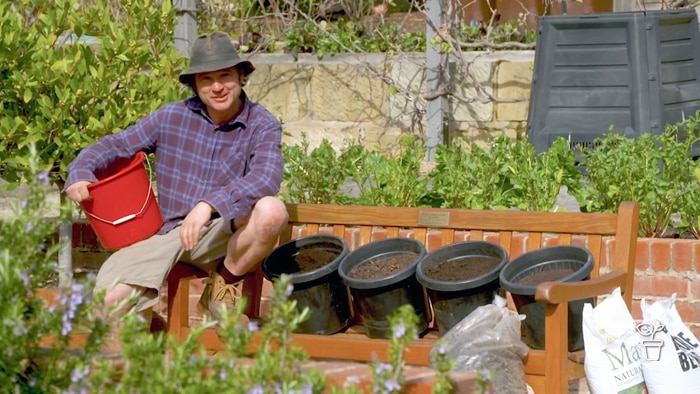 Man in hat with red bucket on lap sitting on edge of the garden bench with black pots filled with manures