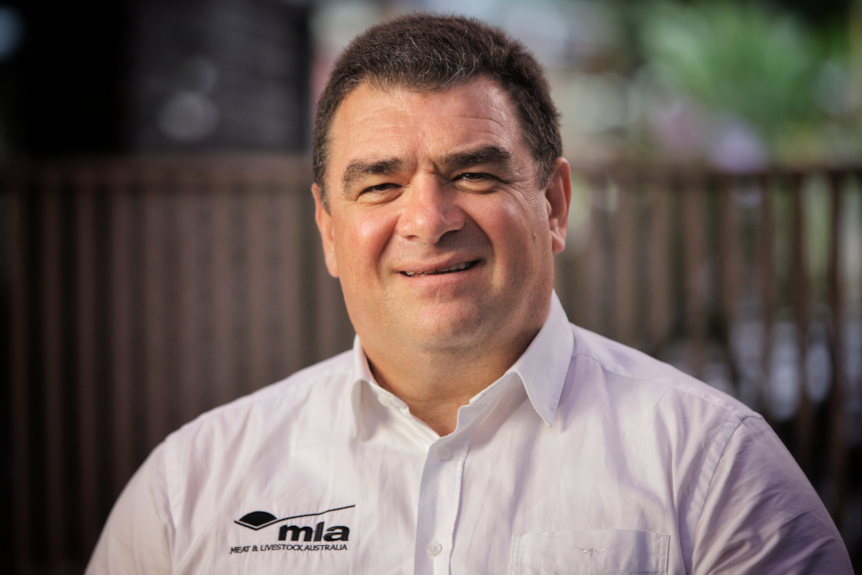 Un homme plus âgé aux cheveux noirs, souriant, portant une chemise à col blanc avec le logo MLA sur sa poitrine droite