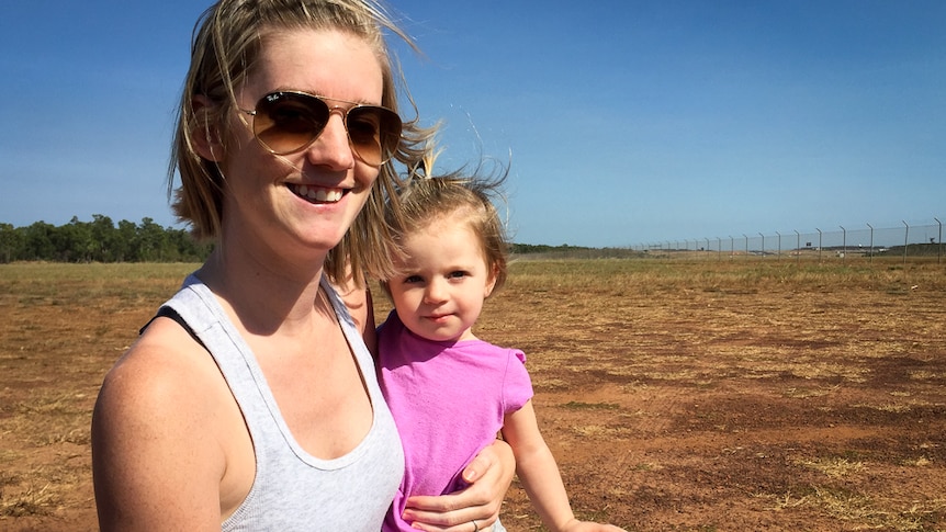 A young mother and daughter stand in a field of dry grass at the end of an airport runway.