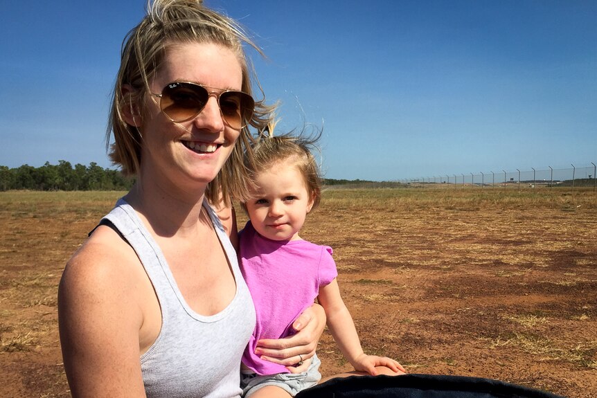 A young mother and daughter stand in a field of dry grass at the end of an airport runway.