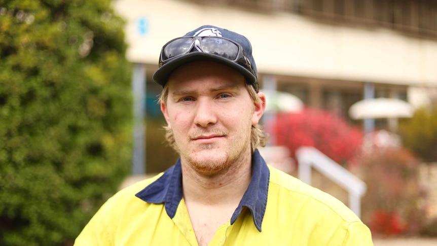Portrait of blonde-haired, blue-eyed man in yellow tradie shirt, with sunglasses and hat on head.