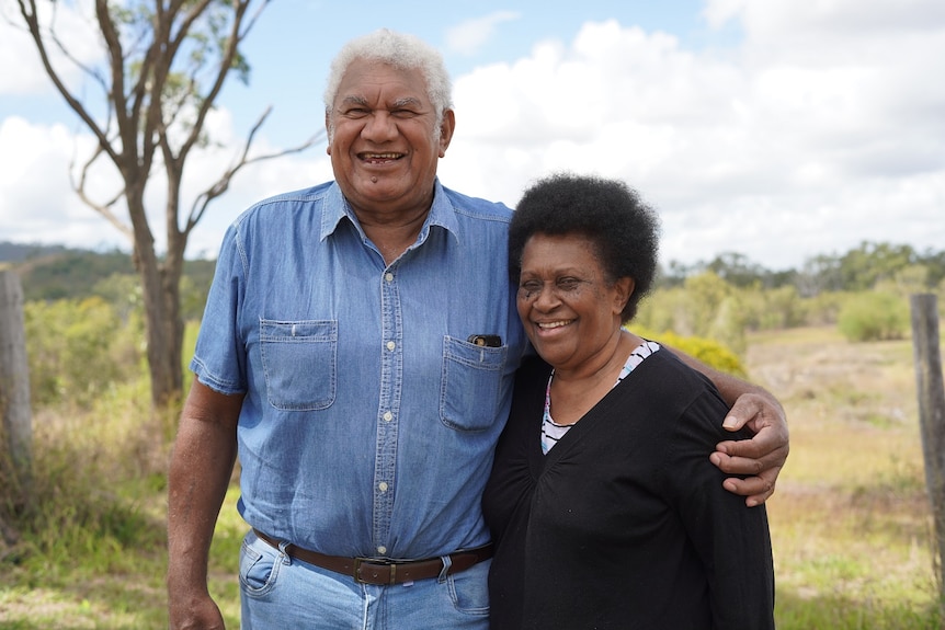 Uncle Butch with his arm around Aunty Vicki, both smiling, grass, trees and blue sky behind.