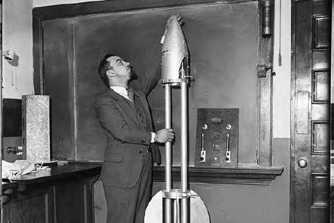 A black and white image of a man in a suit reaching up to the top of a rocket shaped time capsule