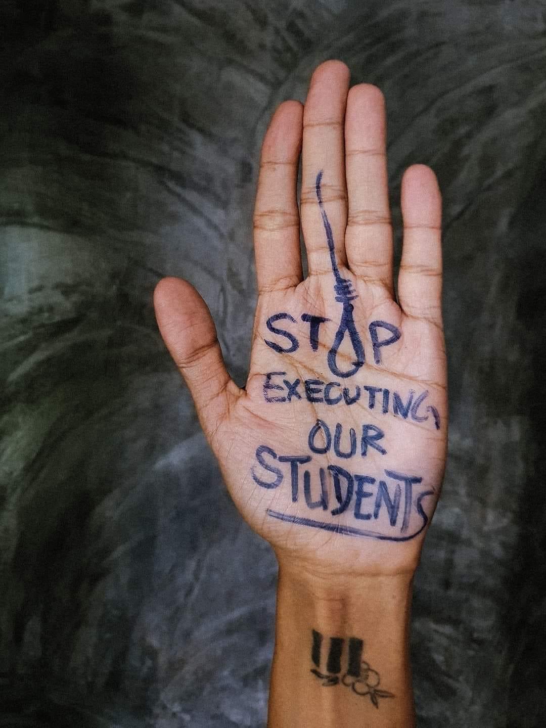 A hand with "stop executing our students" written in pen. The O in STOP is drawn like a noose