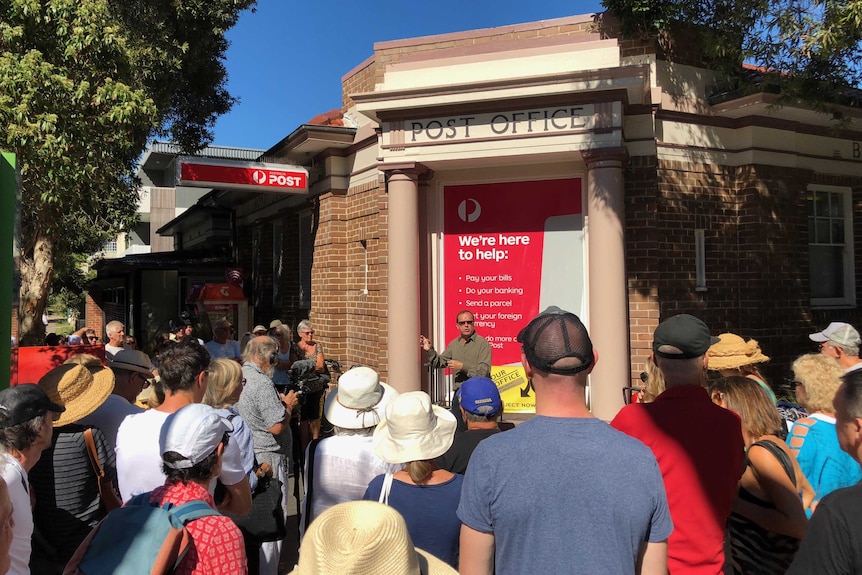 A man speaks to a crowd of people outside a post office building.