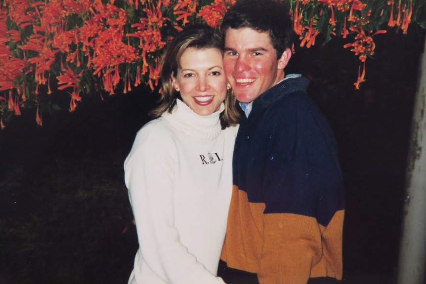 Photo of a young couple- man and woman- standing in front of a vine with red flowers in bloom