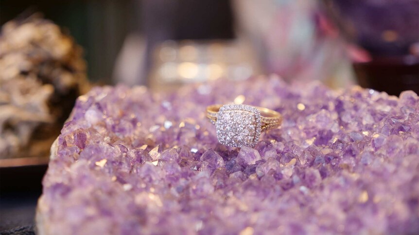 A diamond engagement ring sits on a small bed of purple amethyst crystals