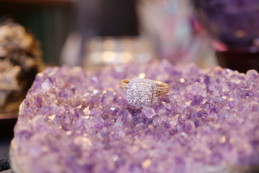 A diamond engagement ring sits on a small bed of purple amethyst crystals