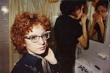 A photograph of a red-haired woman with glasses. Behind her is another woman applying make-up in the mirror.