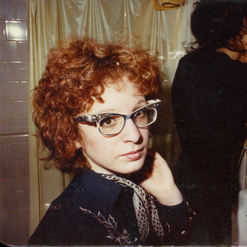 A photograph of a red-haired woman with glasses. Behind her is another woman applying make-up in the mirror.