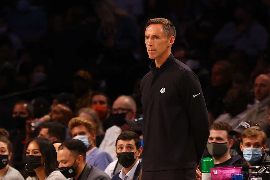 Steve Nash stands with his hands behind his back in front of a crowd of people sitting down
