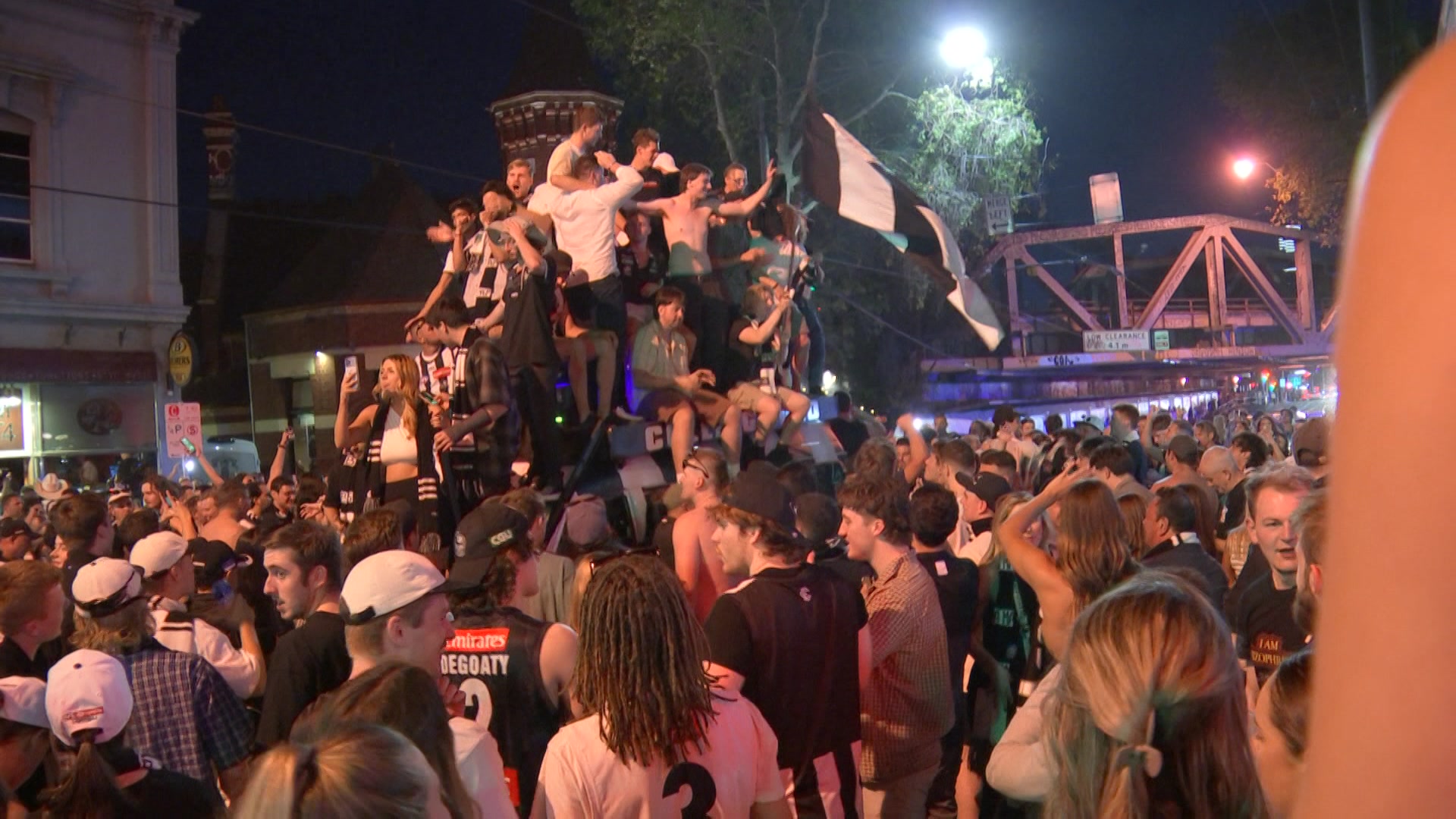 Fans wearing Collingwood clothing crowd around a van that has people piled on top.