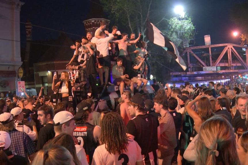 Fans wearing Collingwood clothing crowd around a van that has people piled on top.