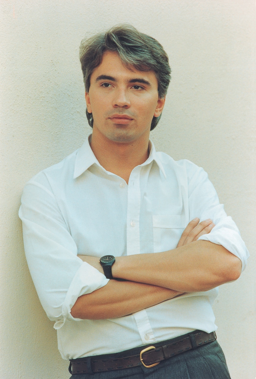 A man in a white shirt leans against a wall with arms crossed.