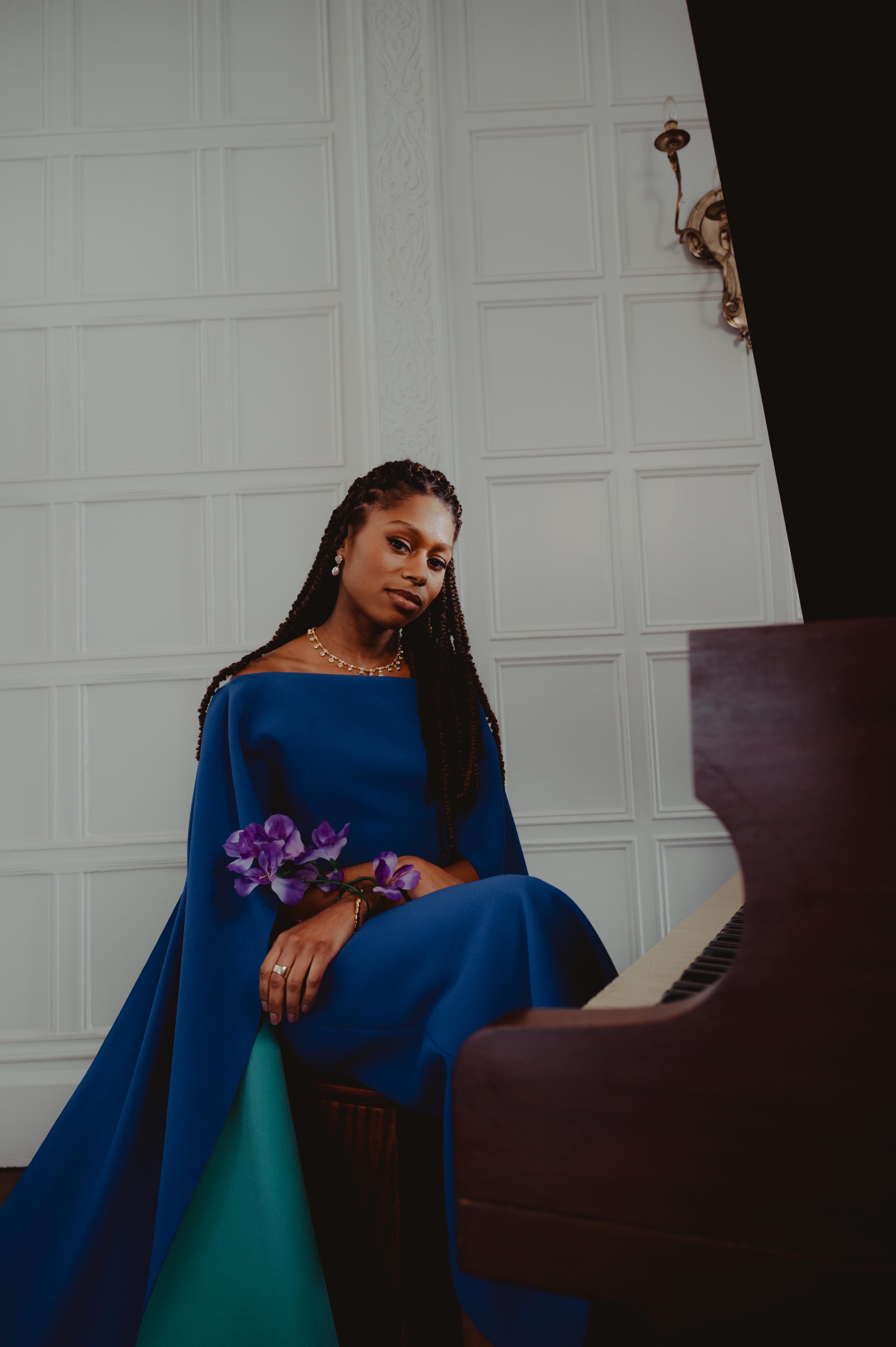 Person in a blue gown seated beside a piano, holding purple flowers against a white paneled wall.