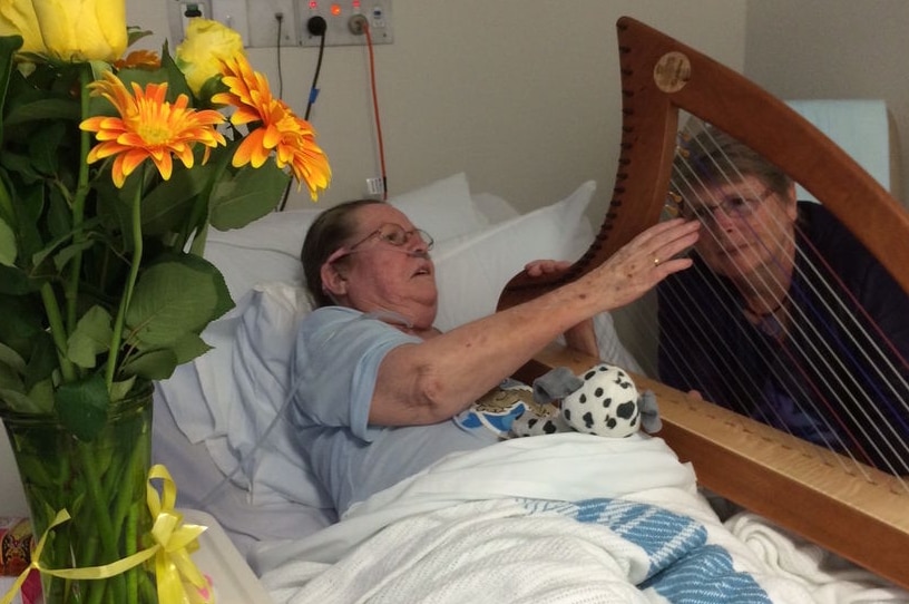 A woman plays a harp to an elderly male patient in a hospital room.