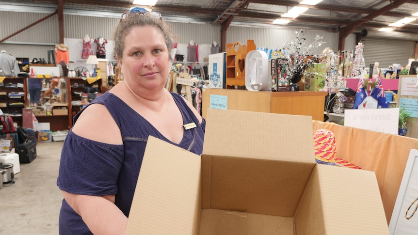 Maria has a neutral expression holding an empty box in front of an isle in the opshop.