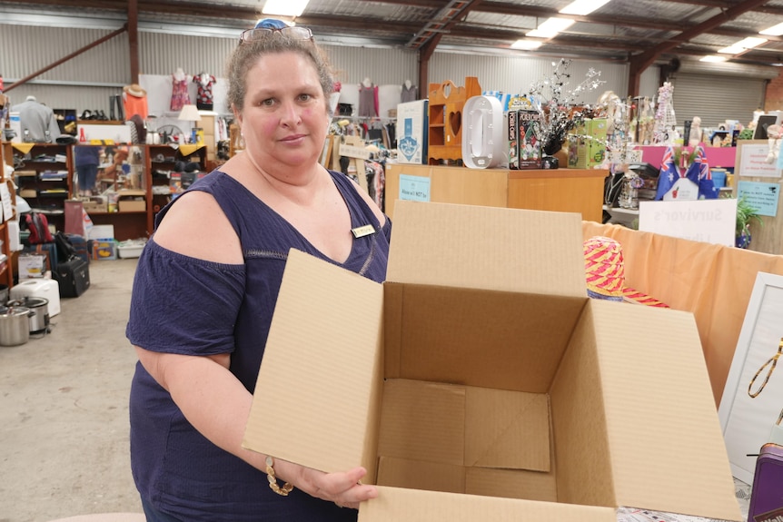 Maria has a neutral expression holding an empty box in front of an isle in the opshop.