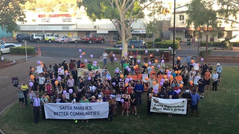 A large group of people stand on a grassy area in front of the main street in Mt Isa, holding lots of purple and orange balloons