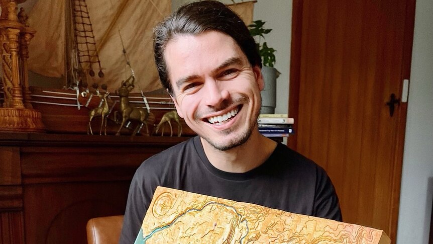 A young man sits in a room holding a map and smiles
