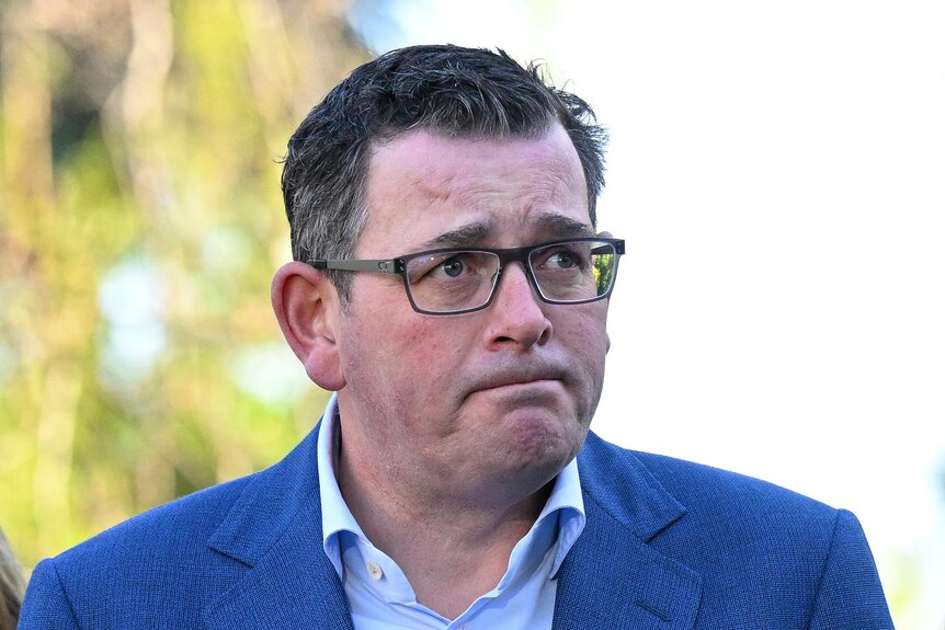 Daniel Andrews wears a blue suit and white shirt and frowns while looking off camera.