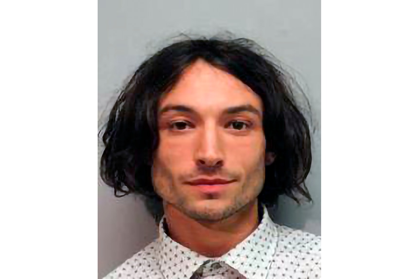 A person with shoulder length dark hair and collared shirt stares into camera for mugshot