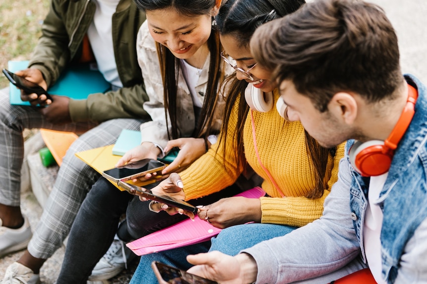 Two teenage girls and a teenage boy sitting together, looking at mobiles phones and smiling