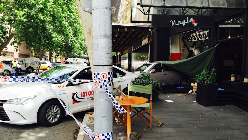 A car inside the smashed shop front of a cafe next to a damaged taxi with police tape surrounding the scene