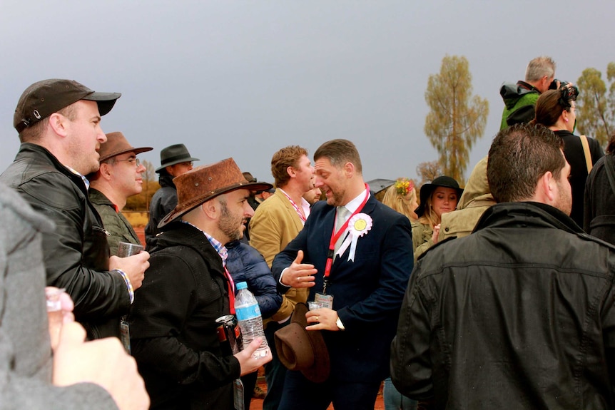 A close shot of a crowd of twenty people watching a camel race. One man in the middle is facing the other way and is laughing.