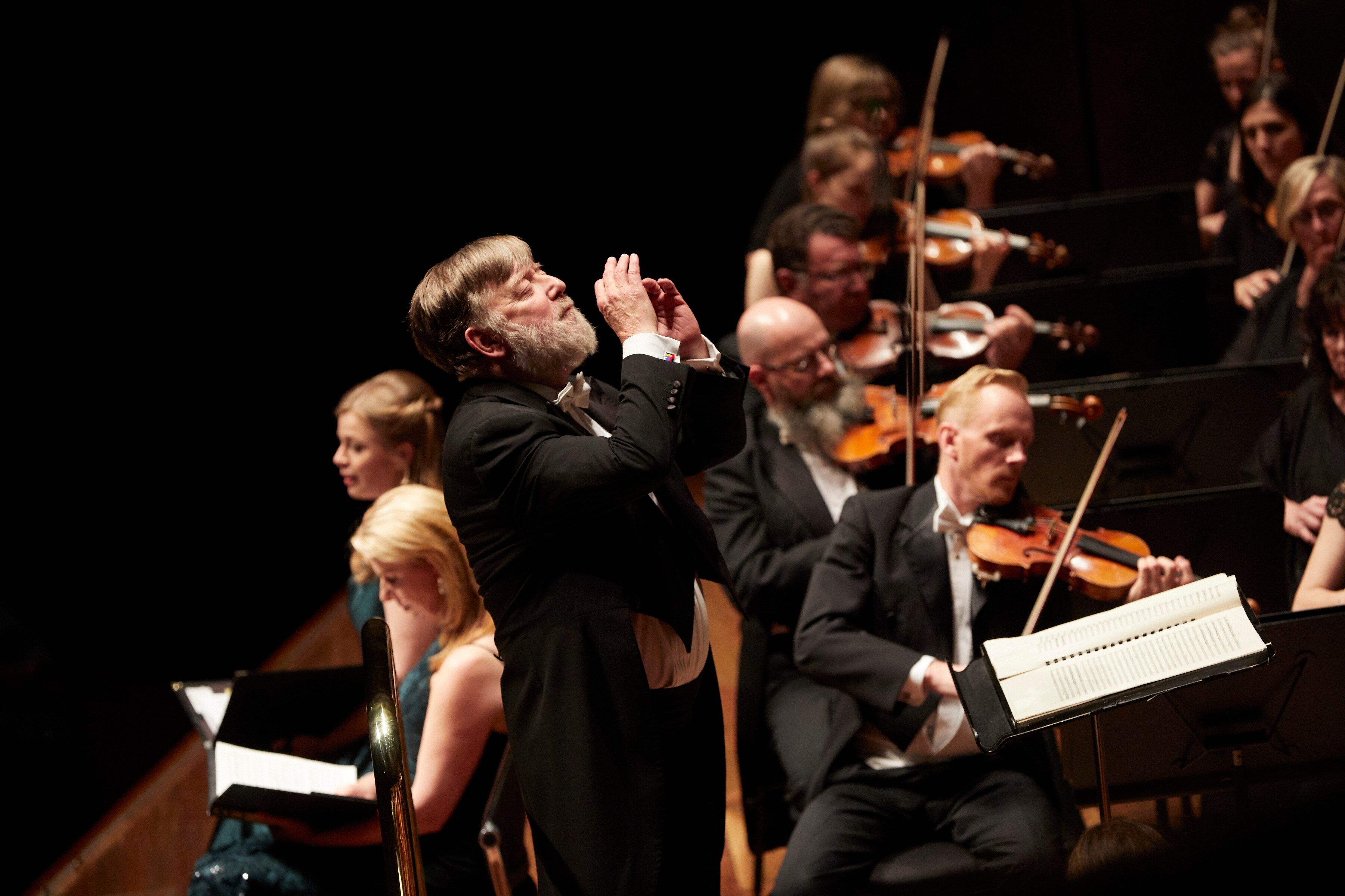Sir Andrew Davis conducts the MSO. He's viewed from the side with his eyes closed and the first violins in the background.
