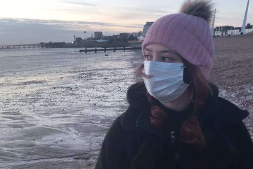 A young woman wearing a pink beanie and a face mask, standing on a beach and looking out towards the ocean.