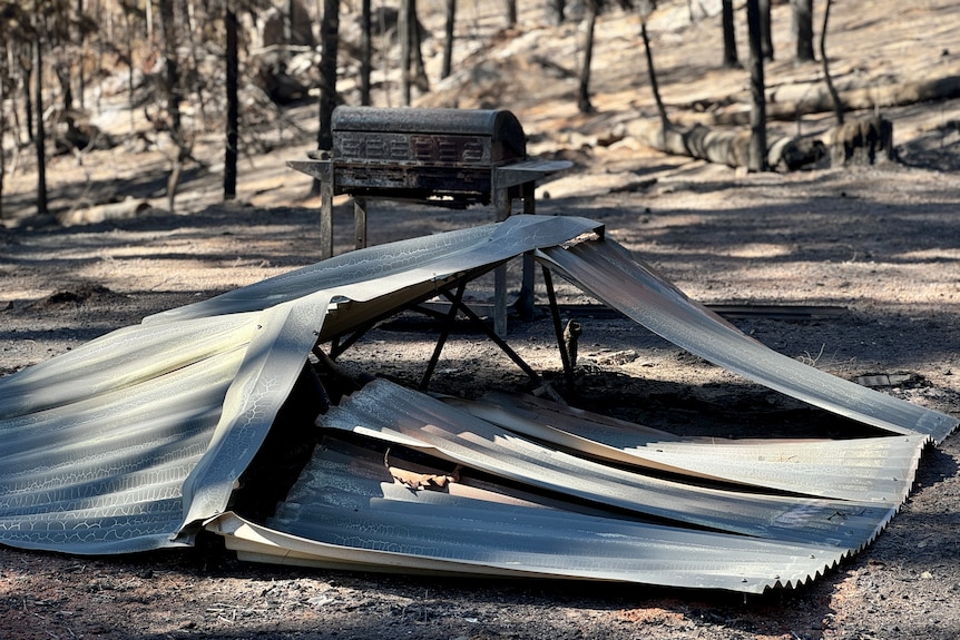 A rectangle of tin roofing lays crumpled in the dirt. A barbecue and black burnt trees stand in the background.